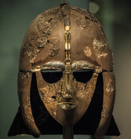 An ancient, weathered metal helmet featuring a decorative gold and silver face mask with intricate engravings, mounted on a stand against a dark background.