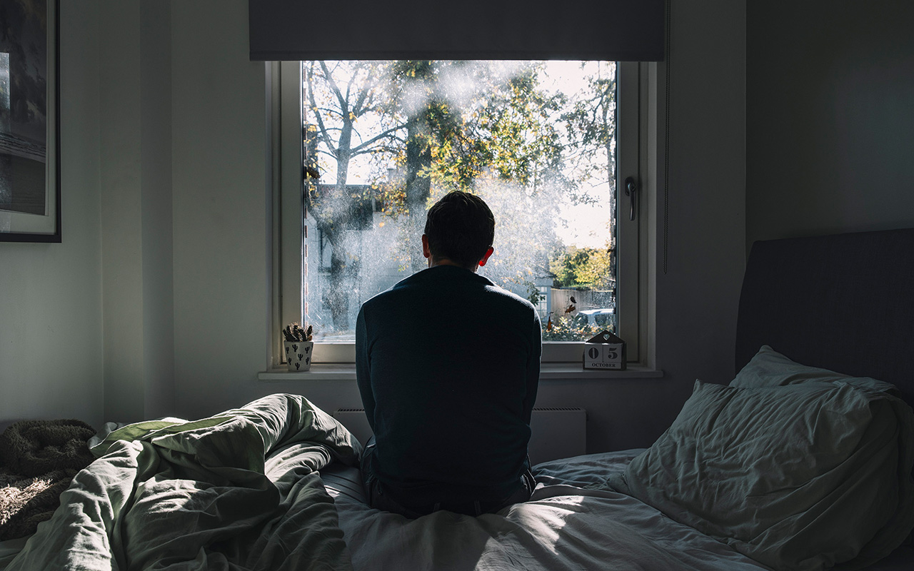 Rear view of an unrecognisable man sitting on his bed looking out of the window 