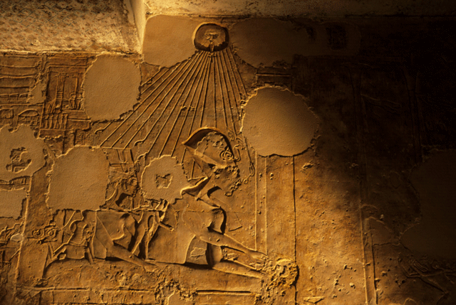 Ancient Egyptian stone relief showing two figures under a sun disk. One figure wears a tall headdress and raises arms, both bask in rays