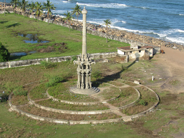 A tall stone monument stands on a circular platform near the shoreline, with palm trees, grassy fields, and ocean waves stretching along the coast behind it.