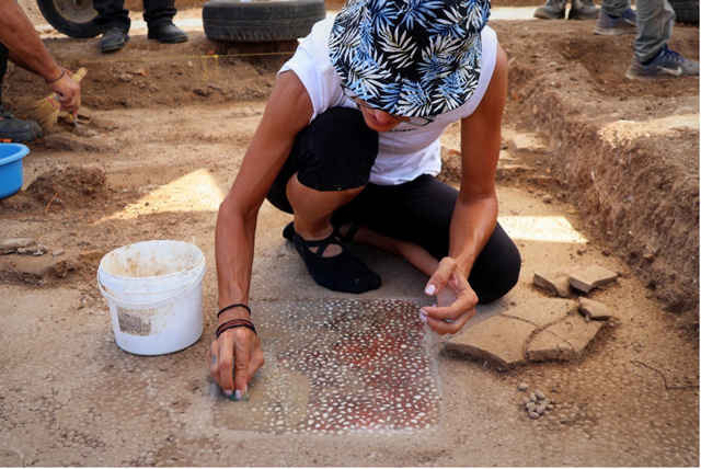 Archaeologists work at an excavation site beneath large white umbrellas, carefully uncovering artifacts and ancient walls inside a series of trenches.