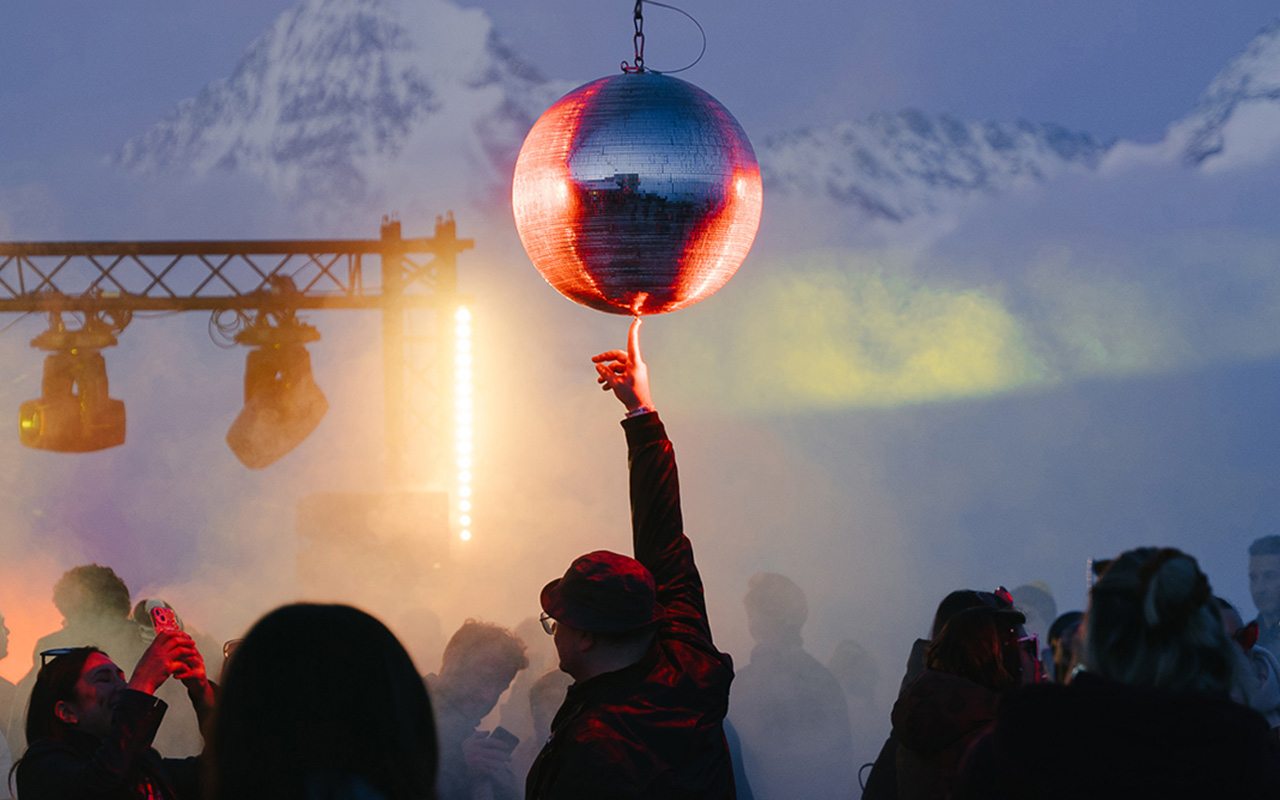 A man points to a mirror ball hanging above a crowd of people at an outdoor party backdropped by snowcapped mountains. 
