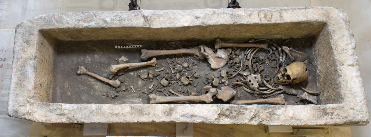 A stone coffin contains the partial skeletal remains of a human, with a skull, ribs, leg bones, and smaller bone fragments arranged within the rectangular stone burial chamber.
