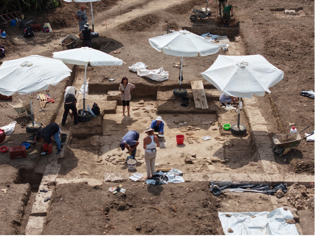An aerial view of an archaeological excavation reveals the stone foundations and layout of ancient buildings exposed beneath the soil.
