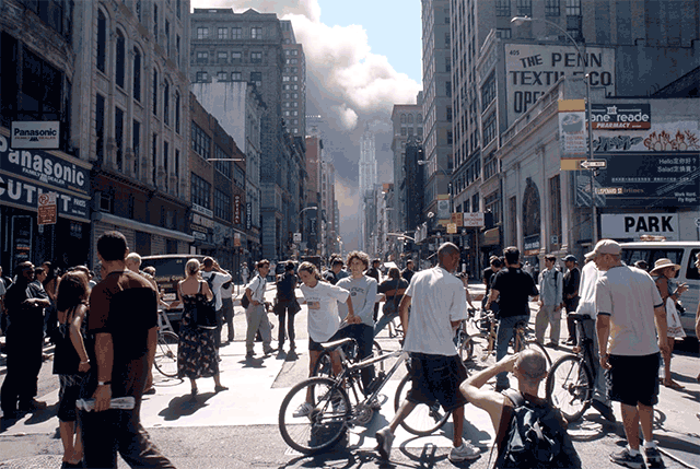 A busy city street with people walking and standing, some with bicycles, and a smoke plume in the distance.