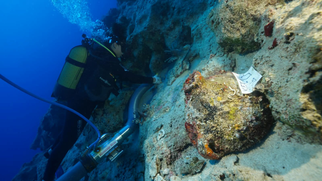 An underwater archaeologist uses excavation equipment to examine a large ancient ceramic vessel resting on the seafloor beside a rocky reef.