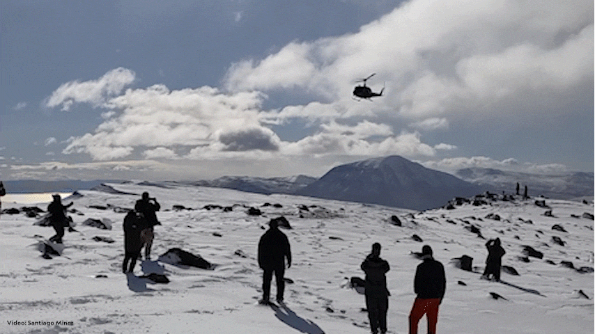 A group standing in winter expedition gear stand on snow covered terrain as they watch an approaching helicopter